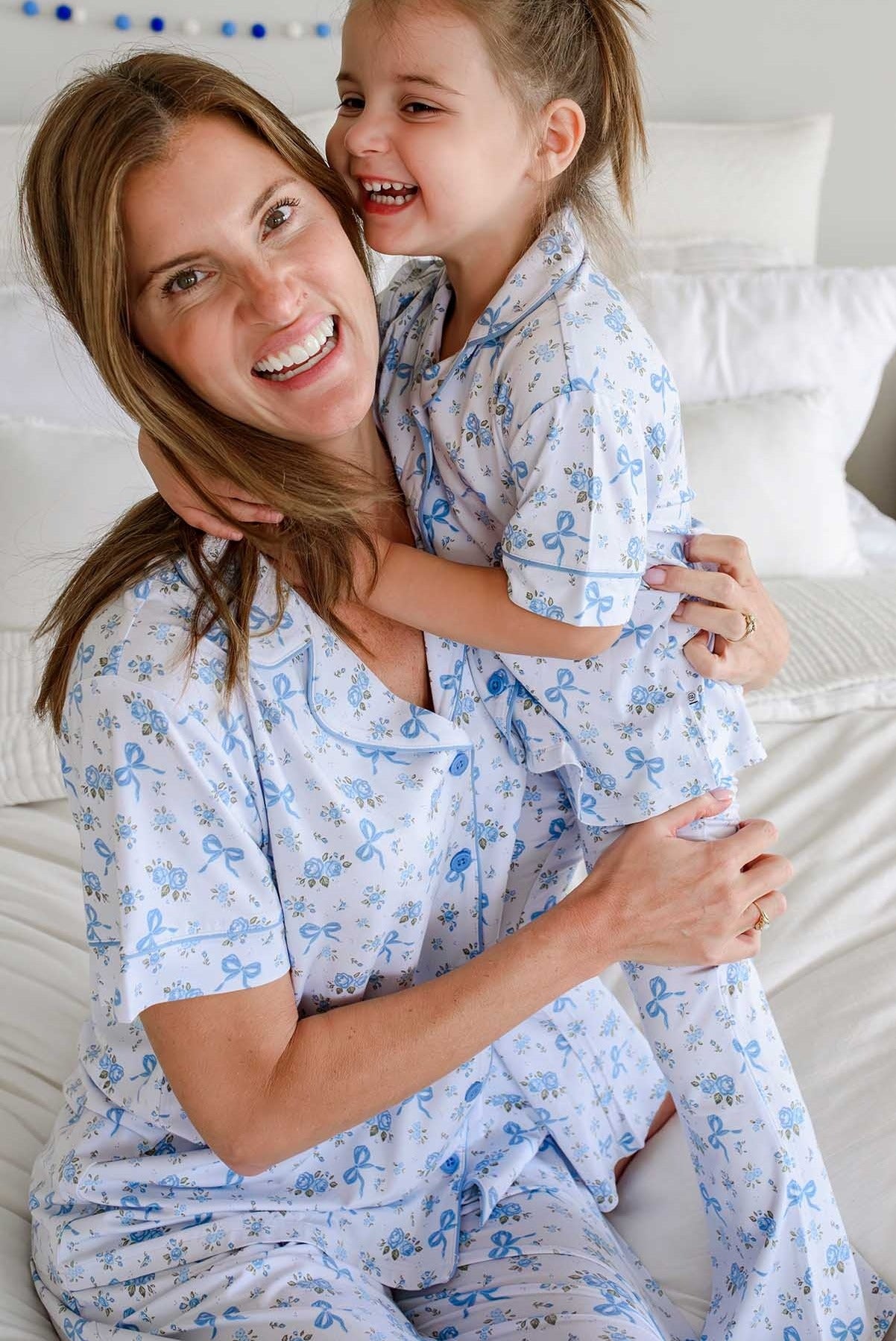 A smiling woman and young girl hugging while wearing matching light blue and white patterned pajamas in a bright bedroom setting.