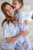 A smiling woman and young girl hugging while wearing matching light blue and white patterned pajamas in a bright bedroom setting.