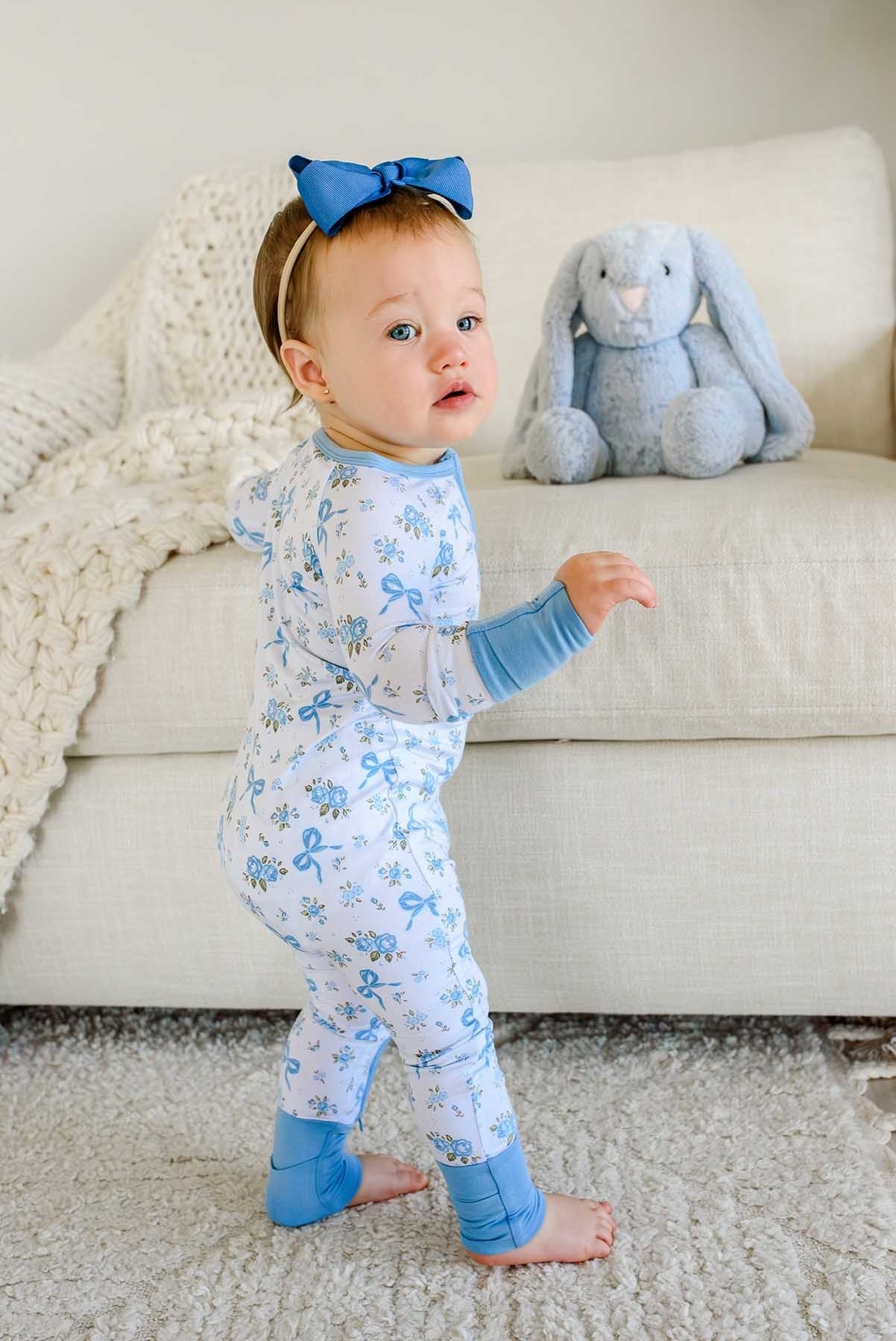 A baby toddler standing on a white shag rug in front of a cream sofa, wearing a light blue and white patterned footed pajama top with blue cuffs and a matching blue bow headband. A gray plush bunny toy sits on the sofa behind the child.