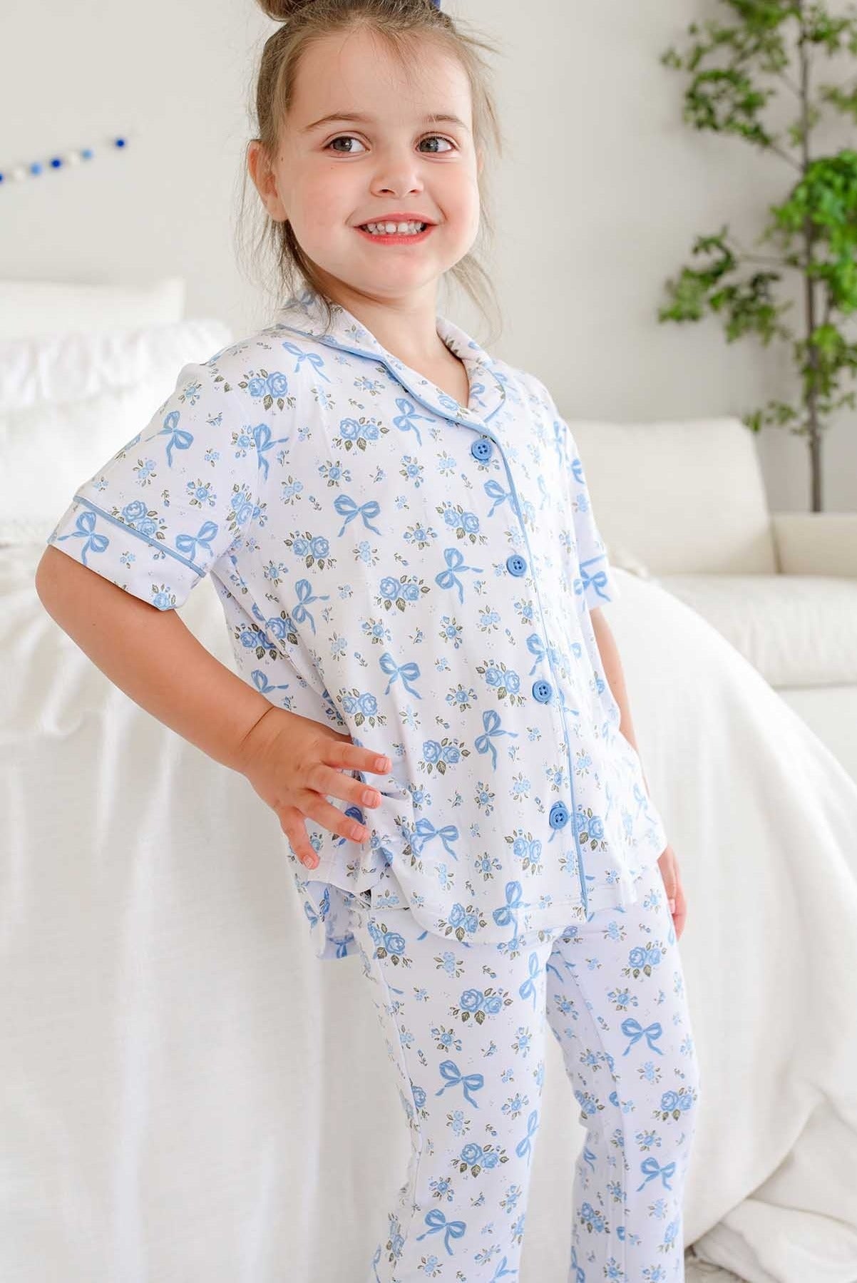 A young girl smiling while wearing a two-piece short-sleeve pajama set with a blue bow and floral pattern on a white background, posed in a bright bedroom setting.