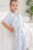 A young girl smiling while wearing a two-piece short-sleeve pajama set with a blue bow and floral pattern on a white background, posed in a bright bedroom setting.