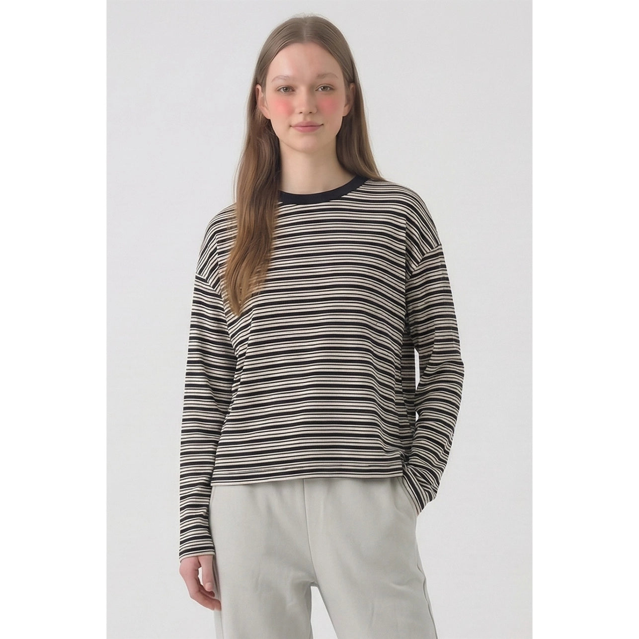A woman modeling a long-sleeve knit top with thin horizontal black, gray, and white stripes, featuring a solid black crew neck, paired with light gray sweatpants against a light gray studio background.
