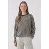 A woman modeling a long-sleeve knit top with thin horizontal black, gray, and white stripes, featuring a solid black crew neck, paired with light gray sweatpants against a light gray studio background.