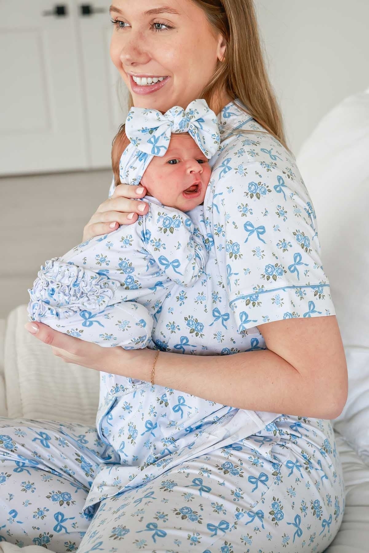 A smiling woman in matching light blue and white patterned pajamas holds a baby wrapped in the same pajama set, with a matching bow headband. The room has a soft neutral background.