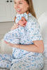 A smiling woman in matching light blue and white patterned pajamas holds a baby wrapped in the same pajama set, with a matching bow headband. The room has a soft neutral background.