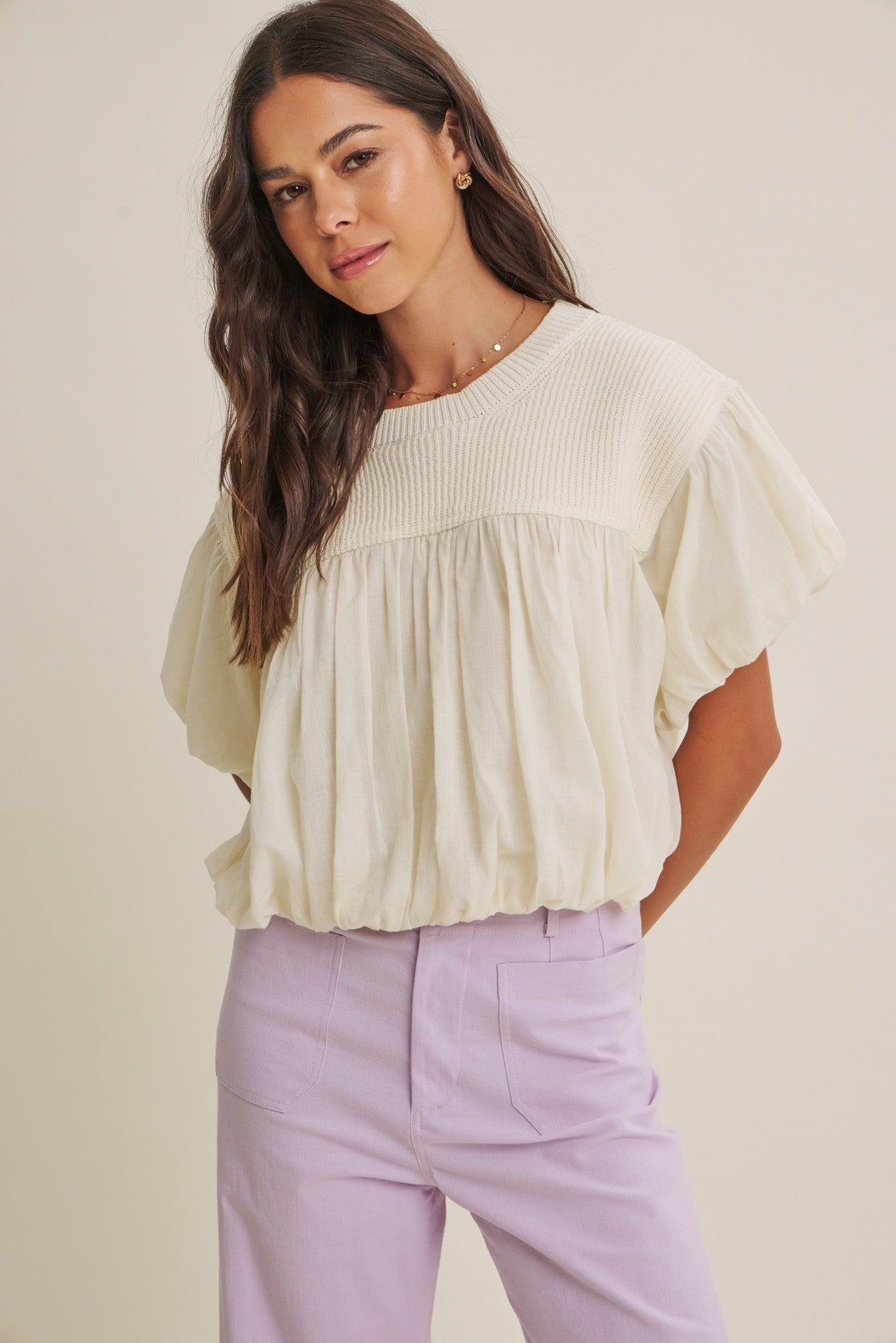 Woman wearing a light-colored blouse and lavender pants against a plain background