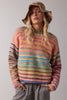 A woman wearing a fuzzy, multicolored striped knit sweater, a brown sequined bucket hat, and gray sweatpants, posing against a neutral light gray studio background.