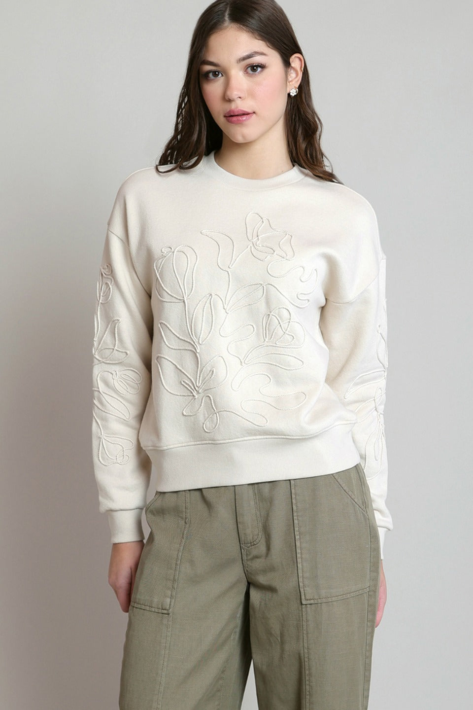 A woman modeling a cream-colored crewneck sweatshirt with tonal floral embroidery on the front and sleeves, paired with olive green cargo pants, against a neutral gray background.