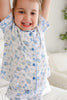 A young child wearing a short-sleeve pajama top with a blue bow and floral print on a white background, smiling with arms raised in a bright indoor setting.