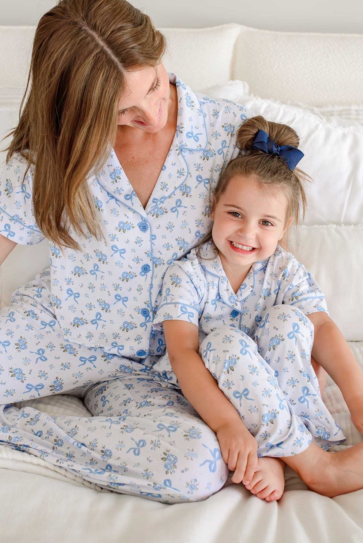 A woman and a young girl sitting together on a white bed, both wearing matching light blue and white floral pajamas.