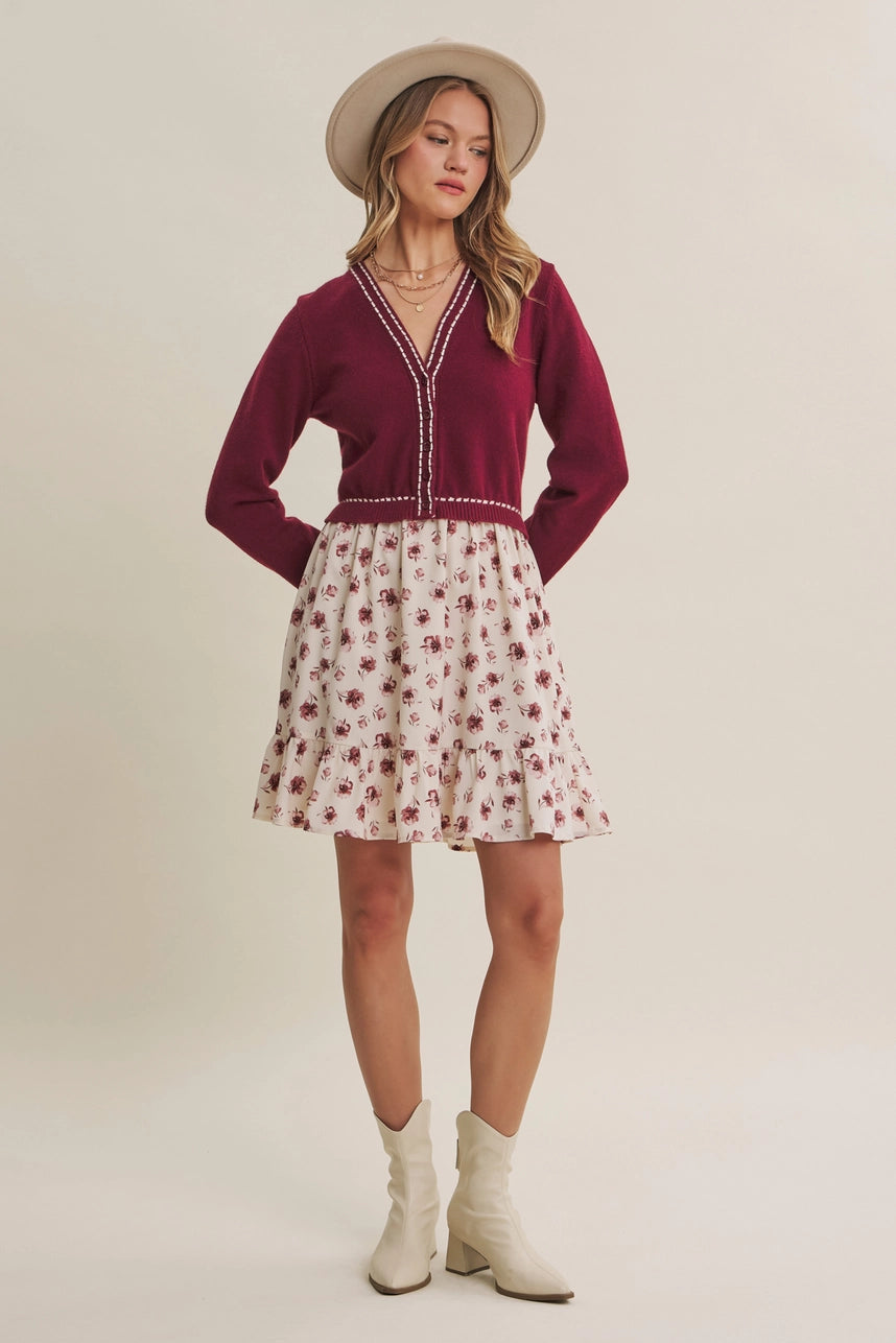 A woman modeling a two-piece outfit consisting of a burgundy knit cardigan with white trim and a cream-colored skirt with a red floral pattern, paired with a beige wide-brim hat and cream ankle boots, against a solid light beige background.