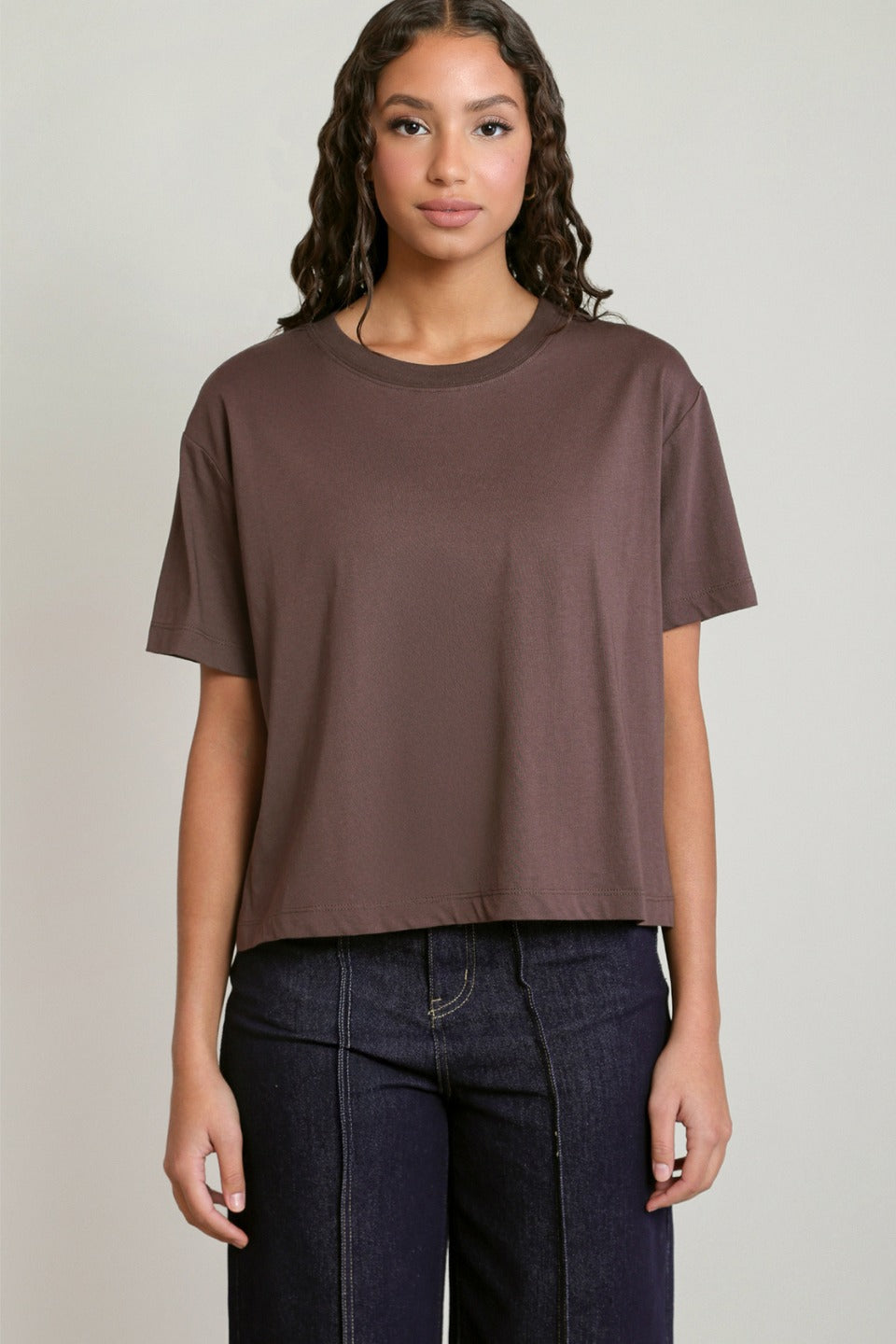A woman modeling a short-sleeve crewneck t-shirt in a muted brownish-purple color, paired with dark blue jeans, against a plain light gray background.