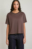 A woman modeling a short-sleeve crewneck t-shirt in a muted brownish-purple color, paired with dark blue jeans, against a plain light gray background.
