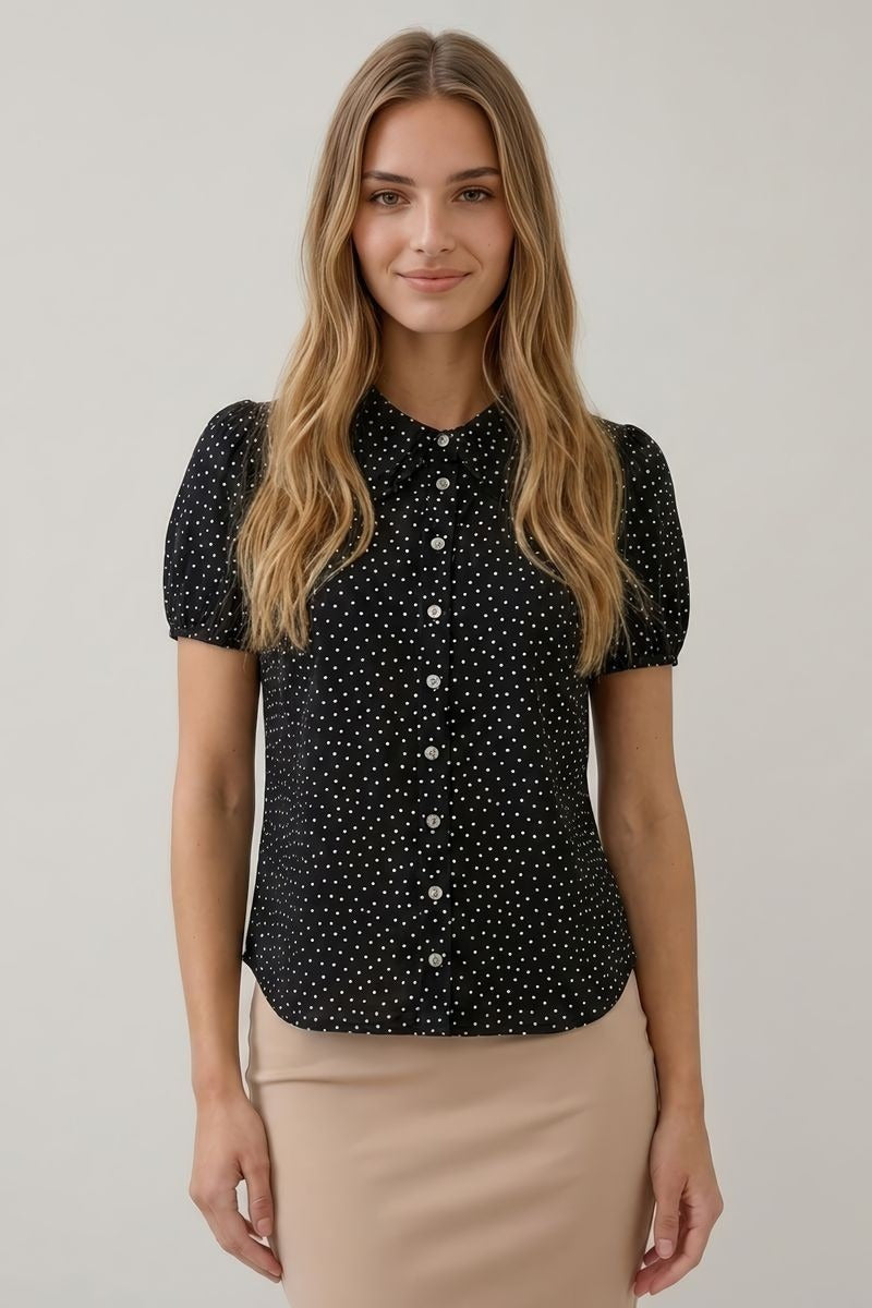 A woman modeling a short-sleeve black blouse with a white polka dot pattern and a button-down front, paired with a beige skirt, against a plain light gray background.