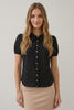 A woman modeling a short-sleeve black blouse with a white polka dot pattern and a button-down front, paired with a beige skirt, against a plain light gray background.