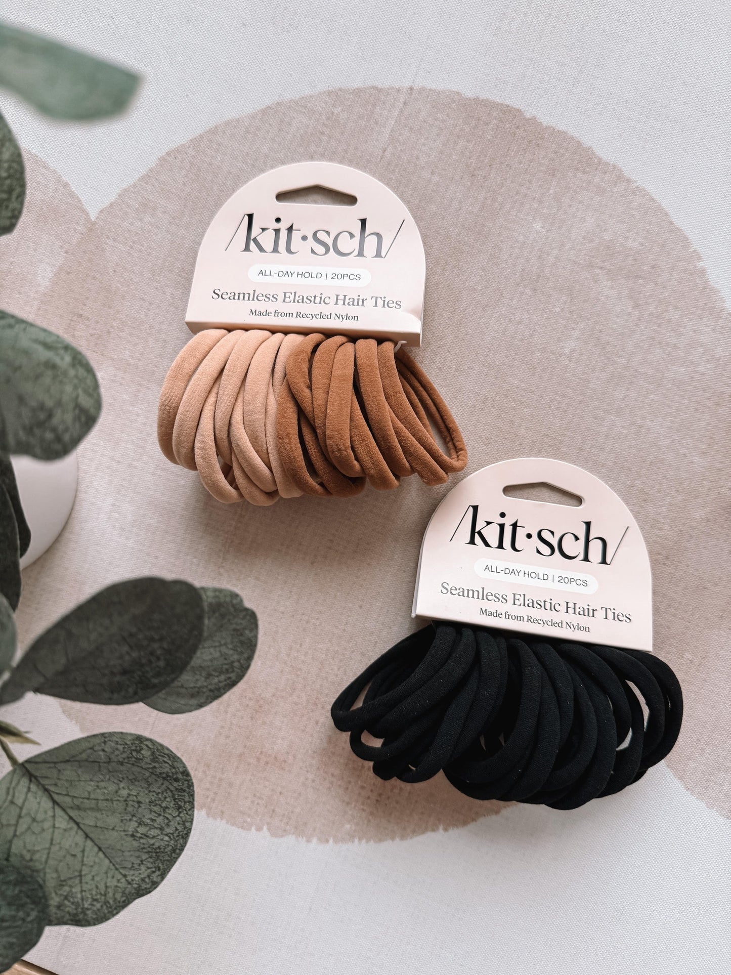 Two packages of kit-sch hair ties, one black and one brown, on a neutral background with green leaves.