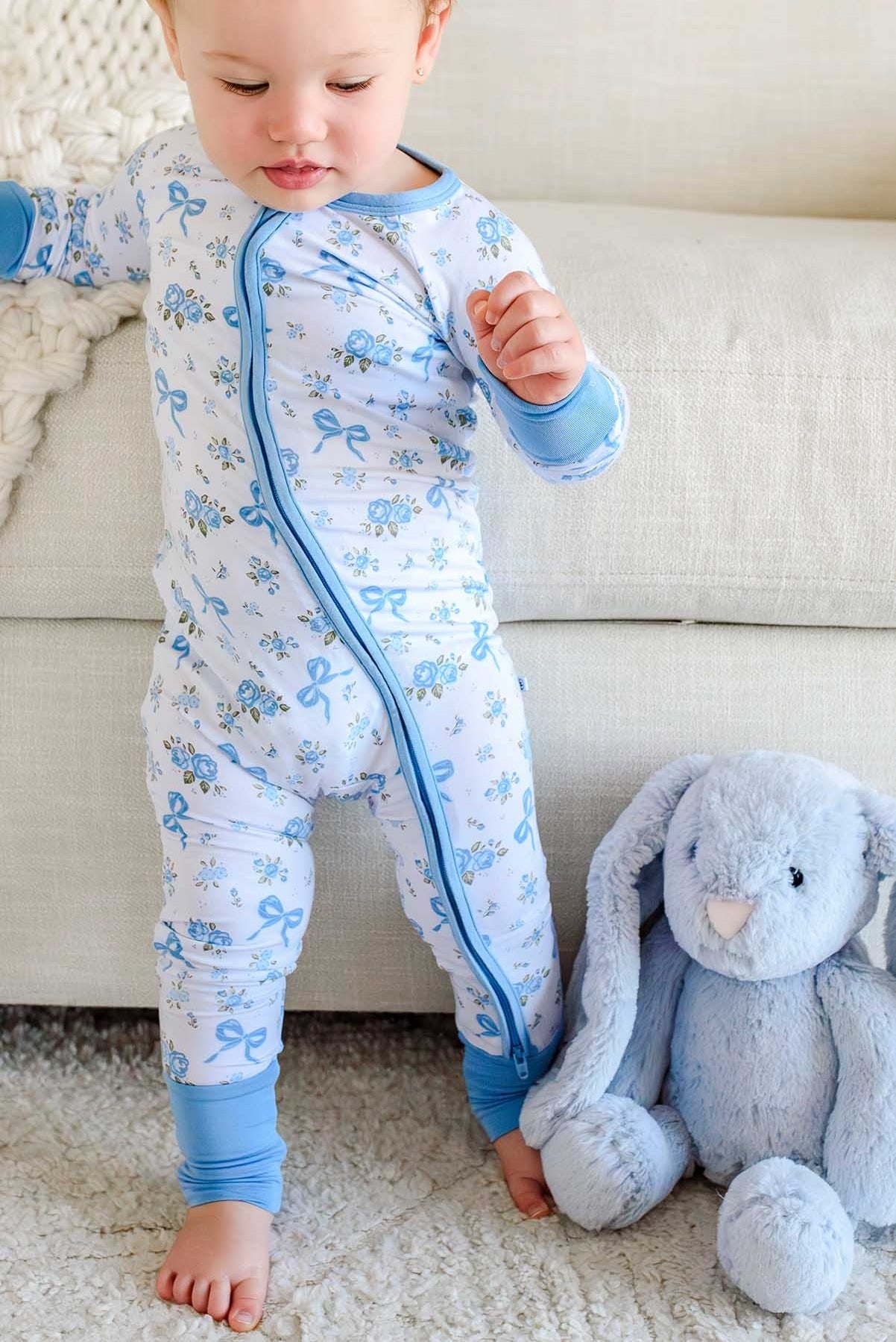 A baby wearing a light blue and white long-sleeve footed sleeper with a blue bow pattern, standing on a white rug next to a gray plush bunny toy in front of a beige sofa.