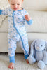 A baby wearing a light blue and white long-sleeve footed sleeper with a blue bow pattern, standing on a white rug next to a gray plush bunny toy in front of a beige sofa.