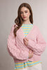 A woman wearing a chunky pink cable-knit cardigan over a yellow and blue striped shirt, photographed against a neutral light gray studio background.