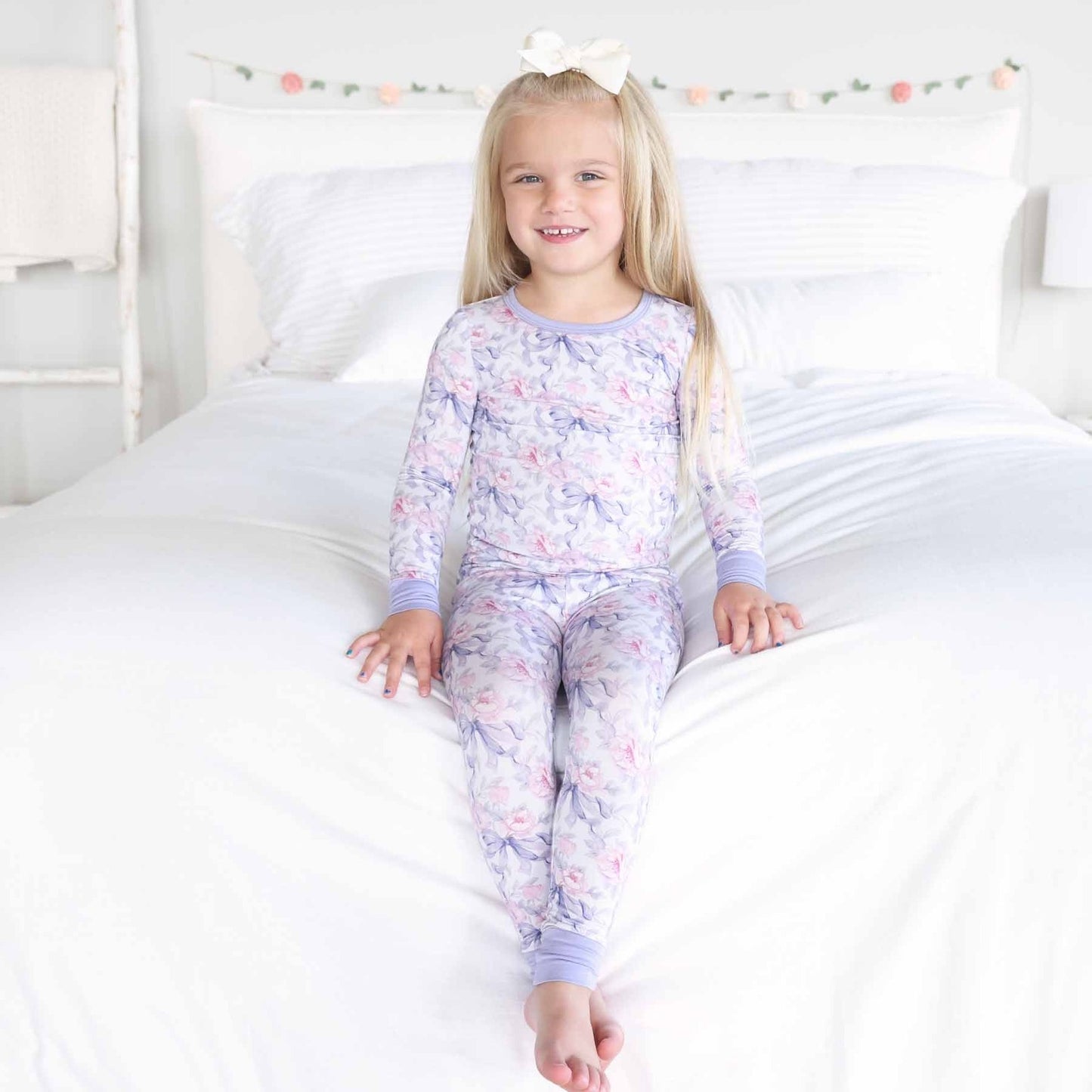 Young girl wearing a floral pajama set sitting on a bed.