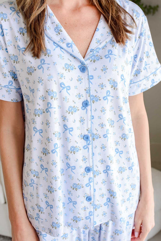 A close-up of a person wearing a short-sleeve pajama top with a light blue and white floral print featuring small bows and flowers, paired with matching pajama pants.