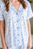 A close-up of a person wearing a short-sleeve pajama top with a light blue and white floral print featuring small bows and flowers, paired with matching pajama pants.