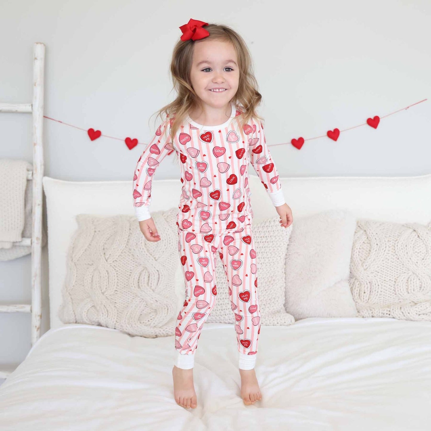 Child wearing a heart-patterned onesie standing on a bed with decorative hearts in the background.