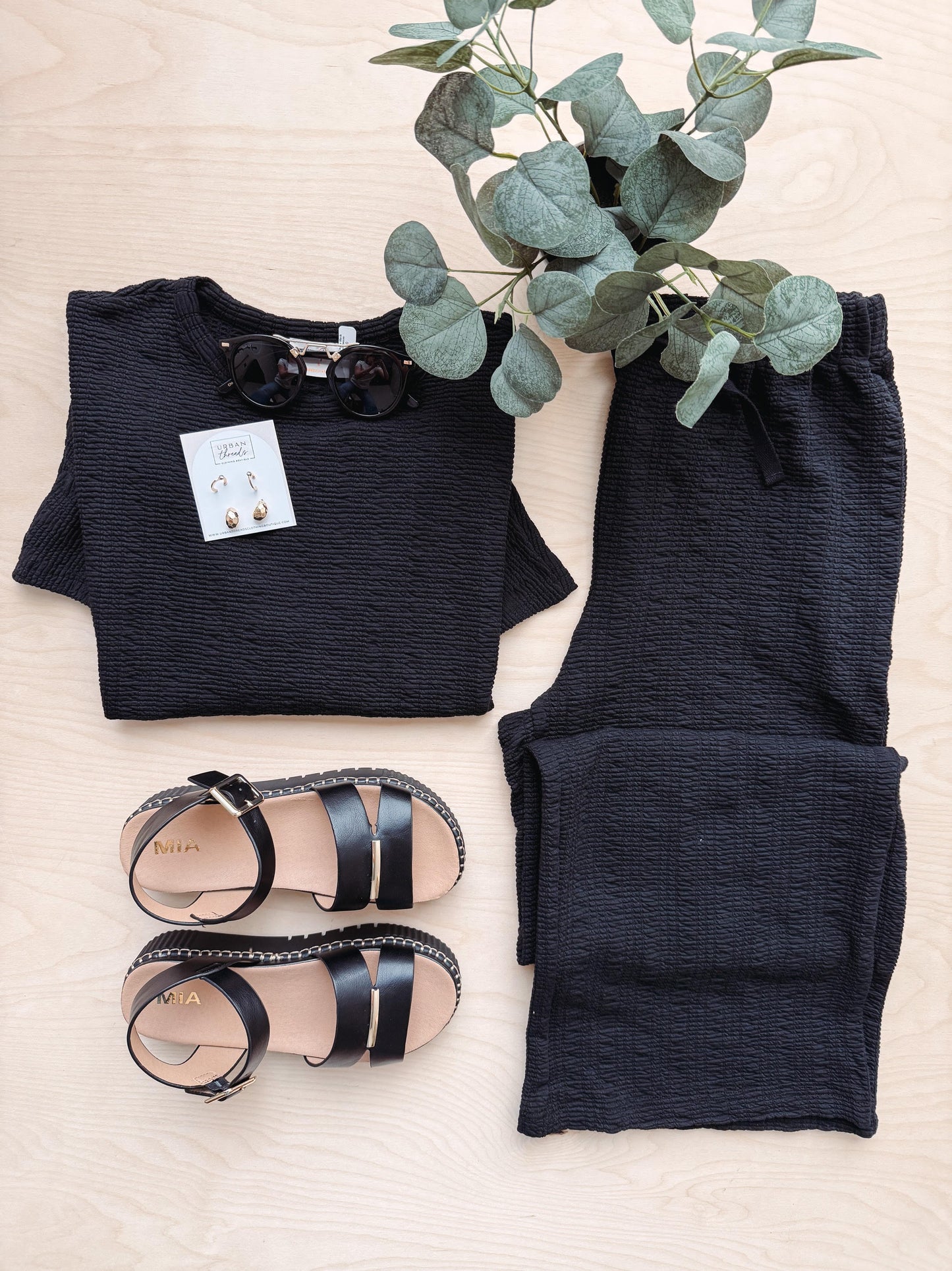 Black outfit with pants, top, sandals, and earrings on a light surface with a plant.