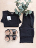 Black outfit with pants, top, sandals, and earrings on a light surface with a plant.