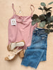 Pink top, blue jeans, sandals, and jewelry on a wooden surface with a plant.