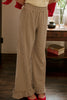 A person wearing wide-leg trousers in a brown and beige gingham pattern, paired with a cream top and red shoes, standing in an indoor room with a wooden floor and furniture in the background.