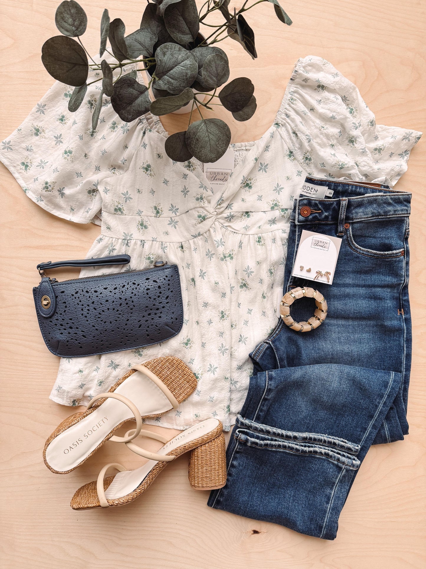 Floral blouse, blue jeans, navy clutch, and sandals on a beige surface with a plant.