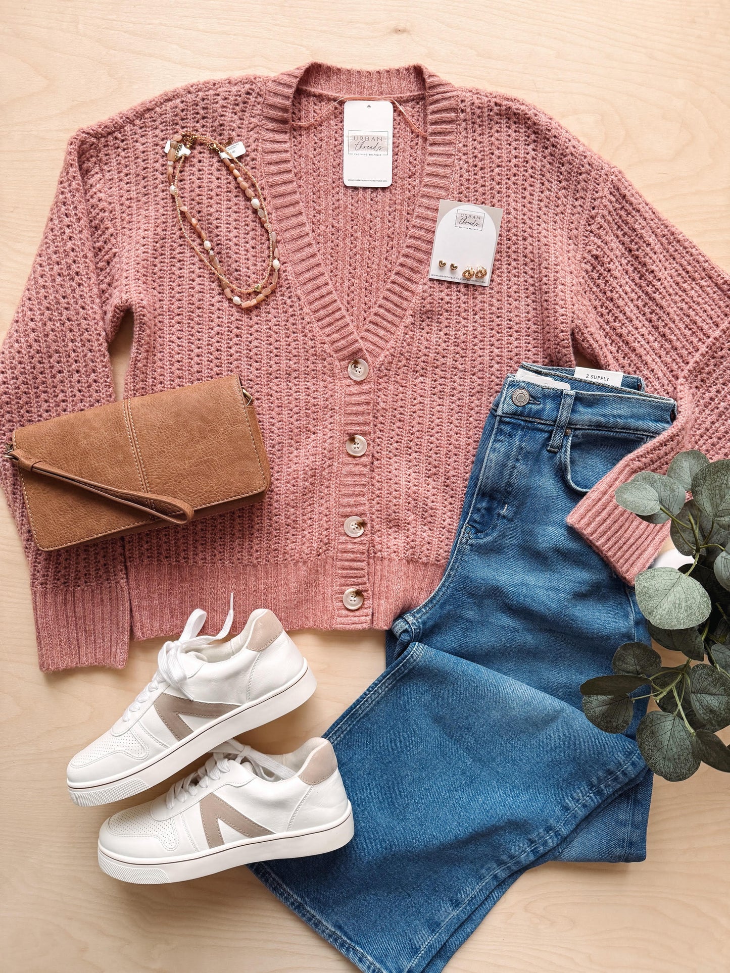 Pink cardigan, white sneakers, blue jeans, brown clutch, and a plant on a wooden surface.