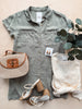 Green shirt, beige bag, sandals, and white baby outfit on a light surface with greenery.
