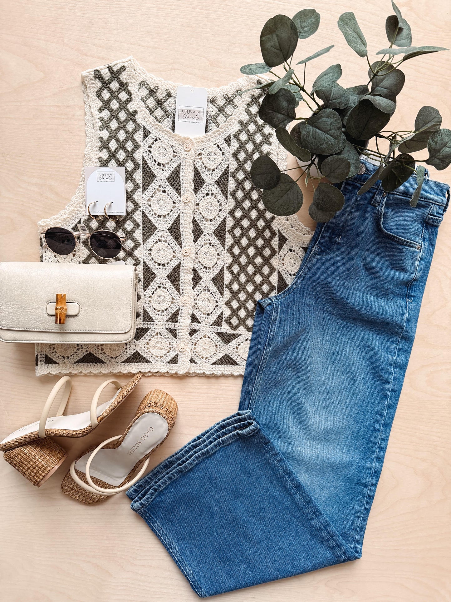 Lace top, blue jeans, white bag, sandals, and sunglasses on a beige surface with greenery.