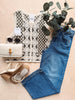 Lace top, blue jeans, white bag, sandals, and sunglasses on a beige surface with greenery.