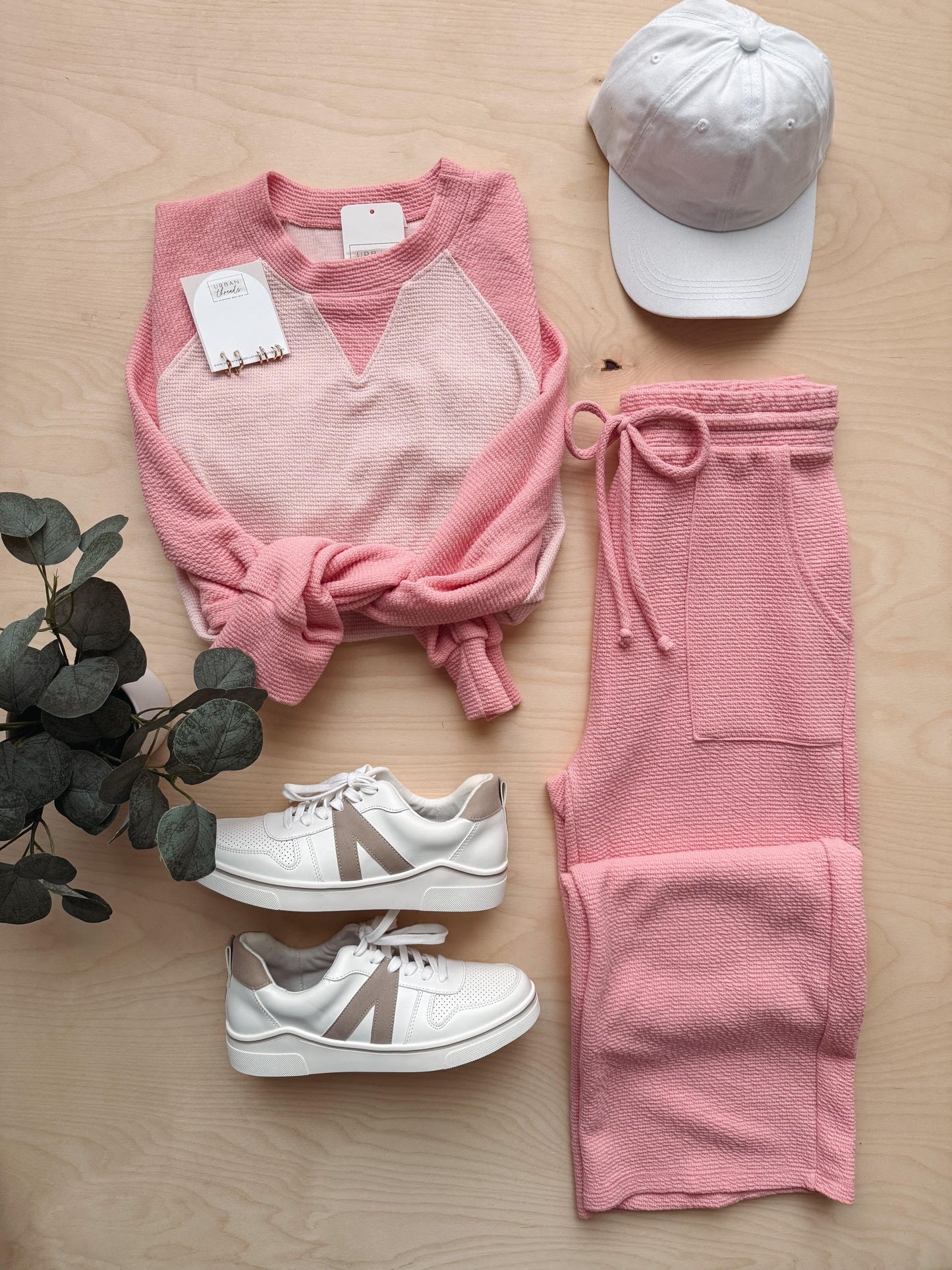 Pink sweatshirt, pink pants, white sneakers, and a cap on a wooden surface with a plant.
