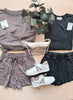 Two outfits laid out on a wooden surface with tags, a plant, and white sneakers.