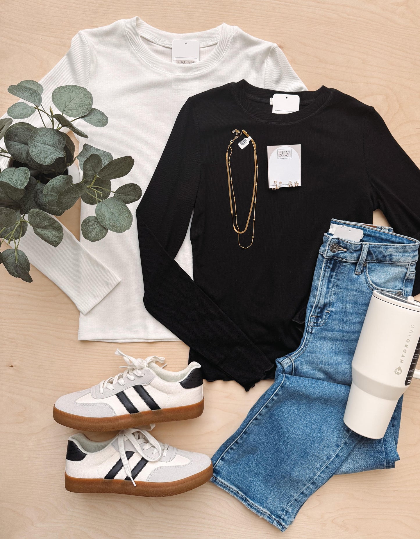 White long-sleeve shirt, black long-sleeve shirt with a necklace, blue jeans, white sneakers, and a white tumbler on a wooden surface.