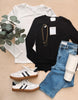 White long-sleeve shirt, black long-sleeve shirt with a necklace, blue jeans, white sneakers, and a white tumbler on a wooden surface.
