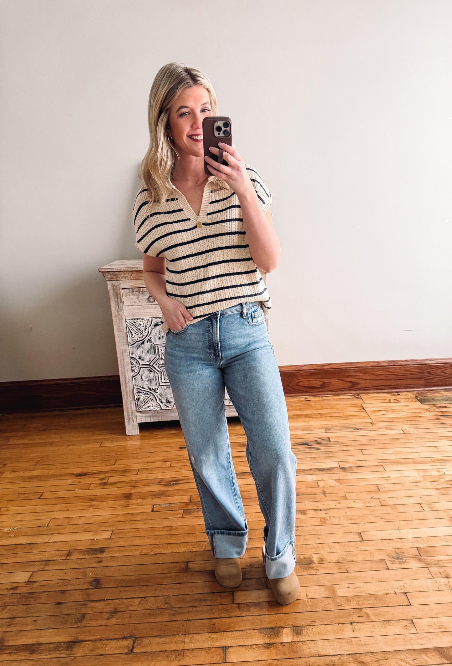 Woman taking a mirror selfie wearing a striped shirt and jeans in a room with wooden flooring.
