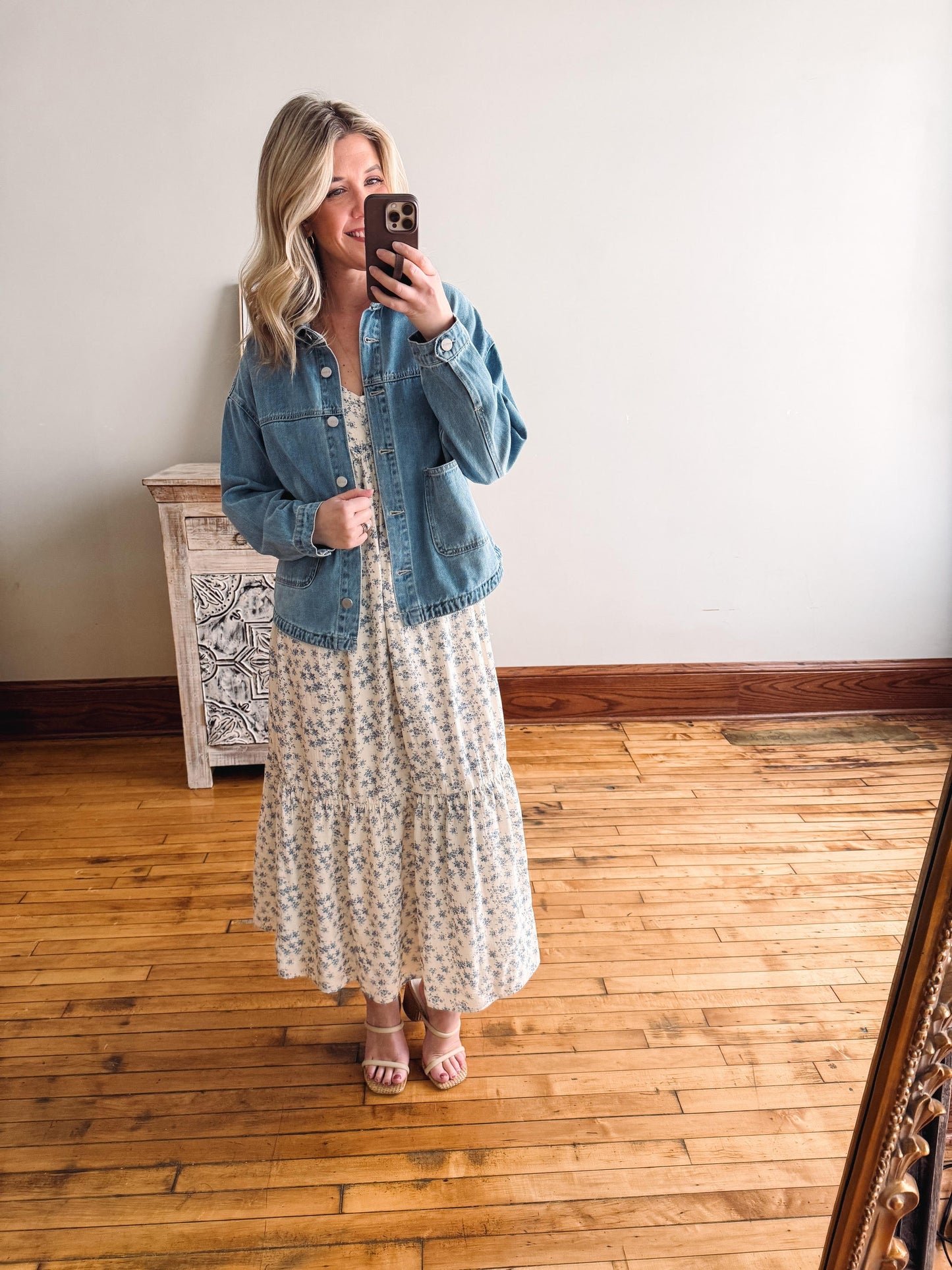Woman taking a mirror selfie wearing a floral dress and denim jacket in a room with wooden flooring.