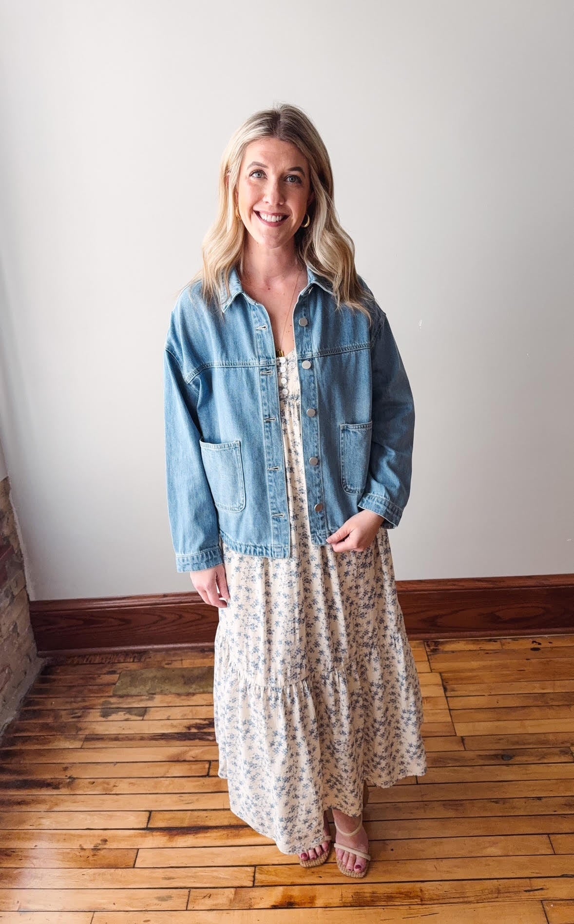 Woman wearing a denim jacket over a floral dress on a wooden floor.