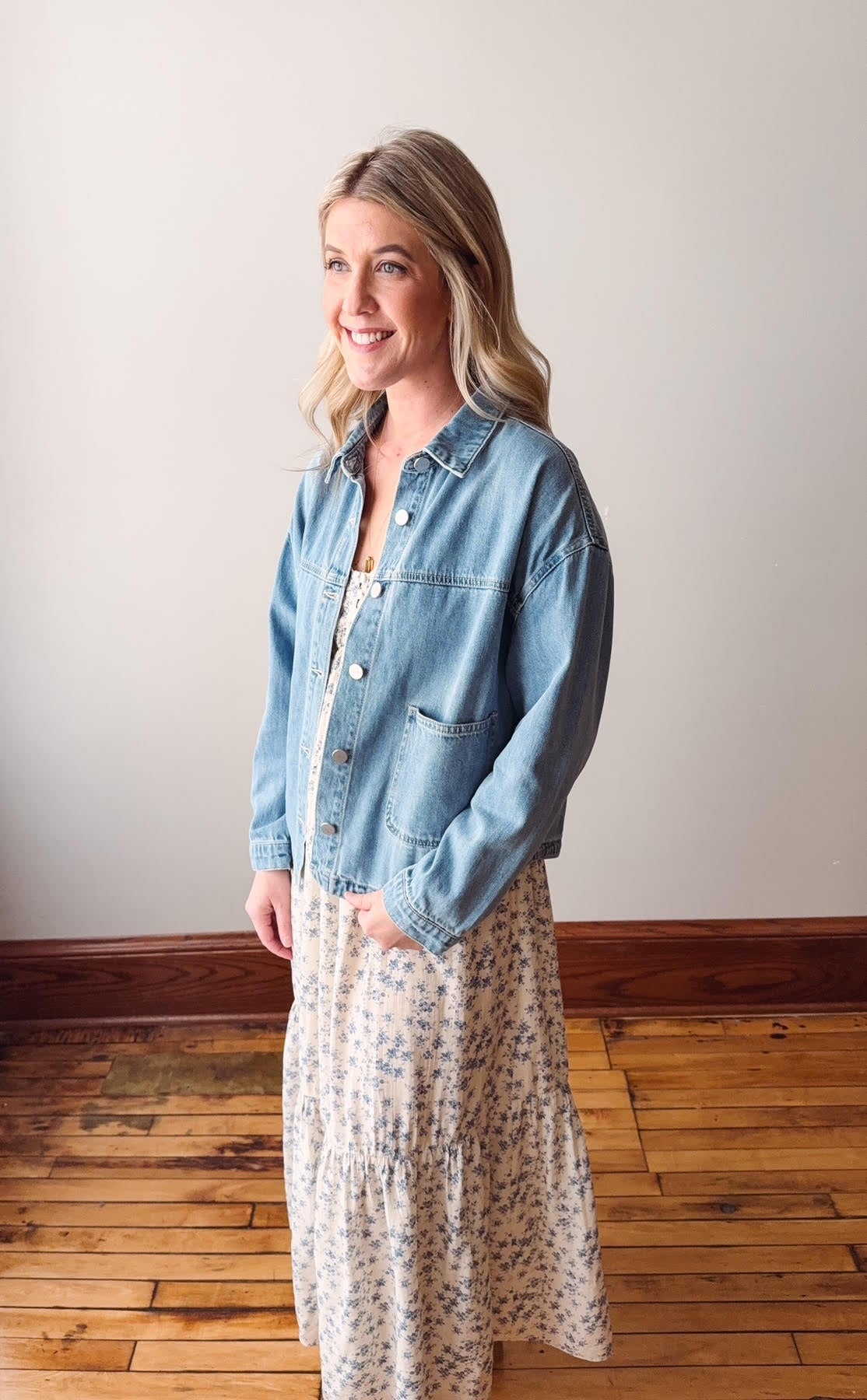 Woman wearing a denim jacket and floral dress standing on a wooden floor.