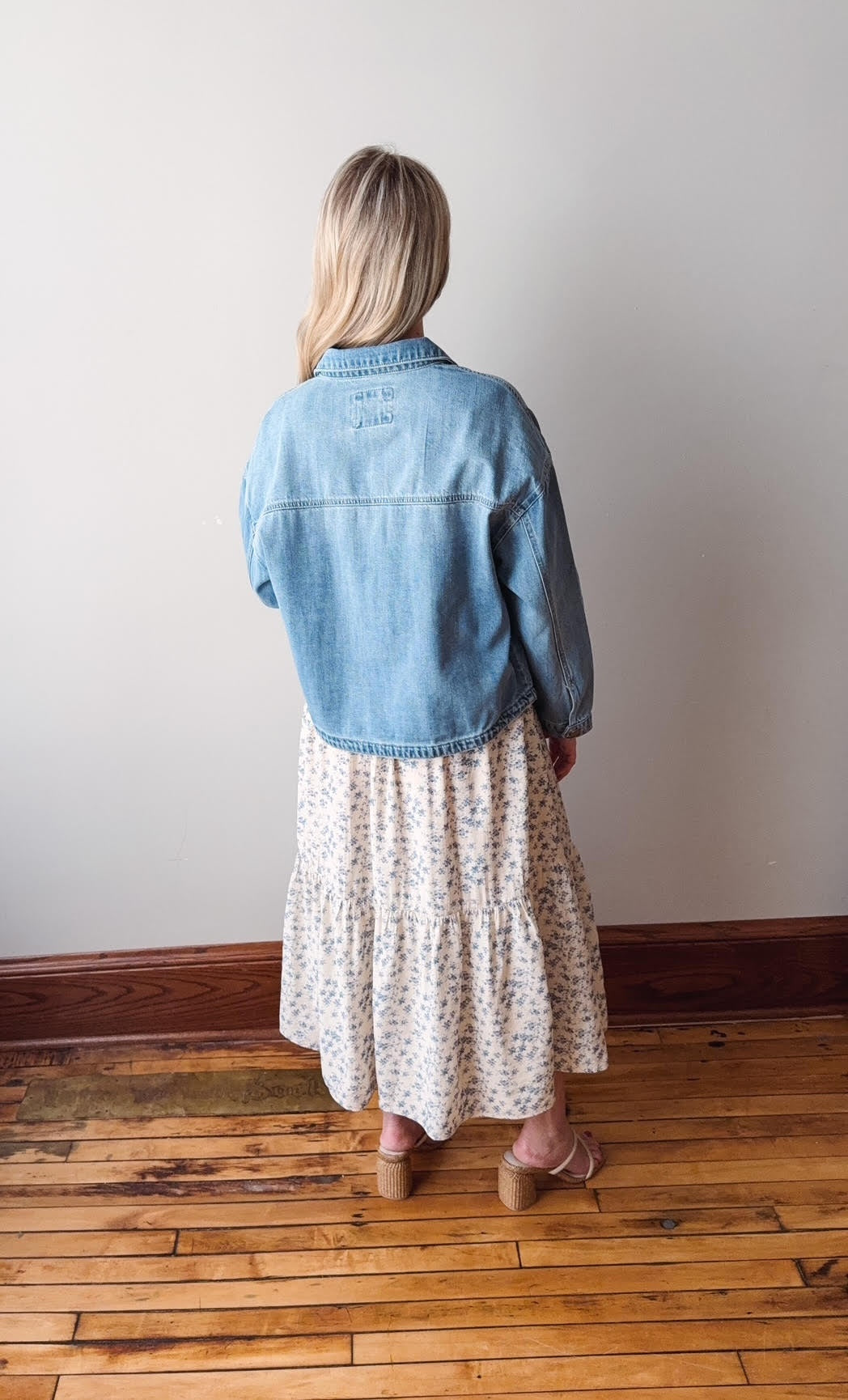 Person wearing a denim jacket and floral dress standing on a wooden floor.