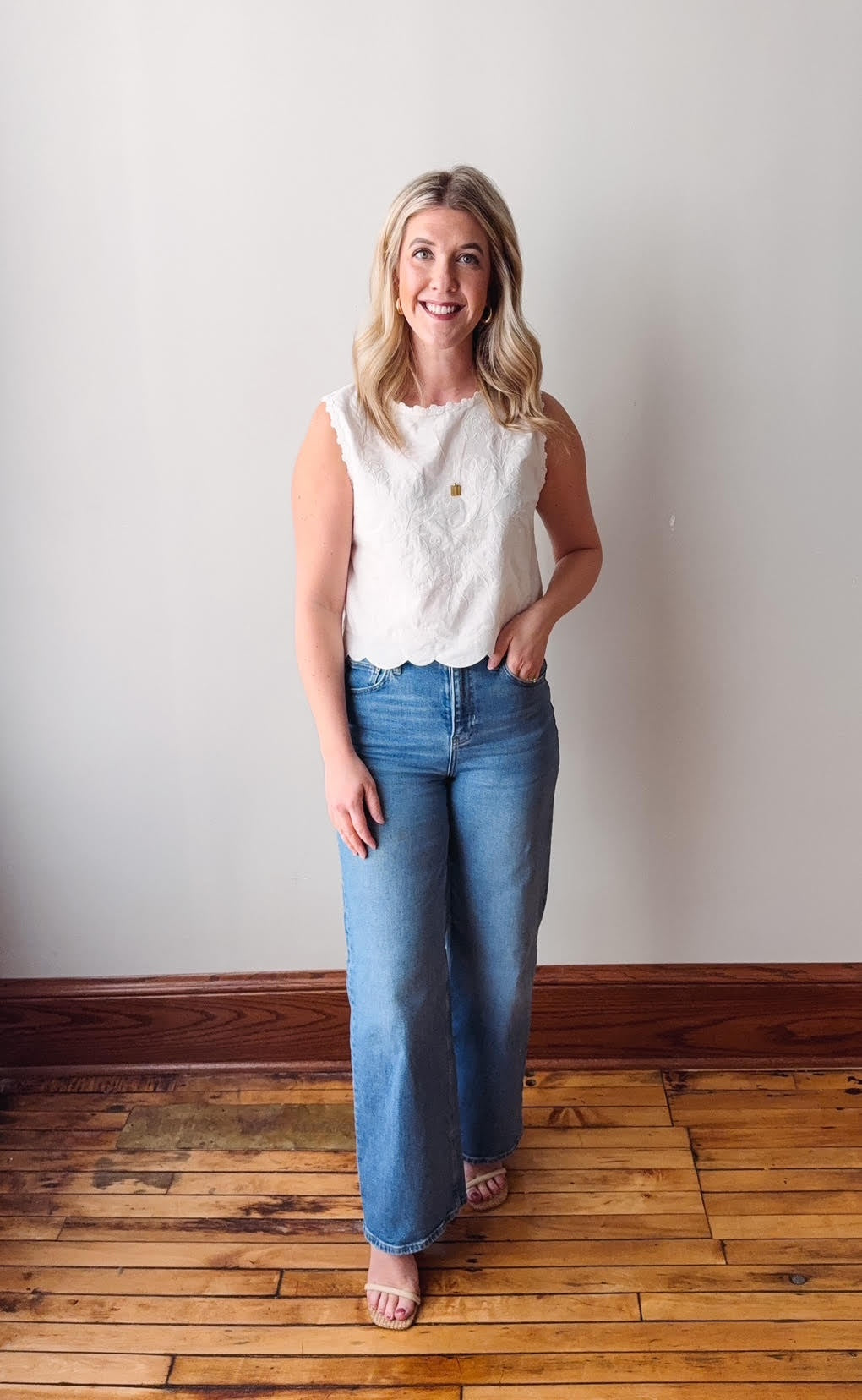 Woman wearing a white sleeveless top and blue jeans standing on a wooden floor with a plain wall background.