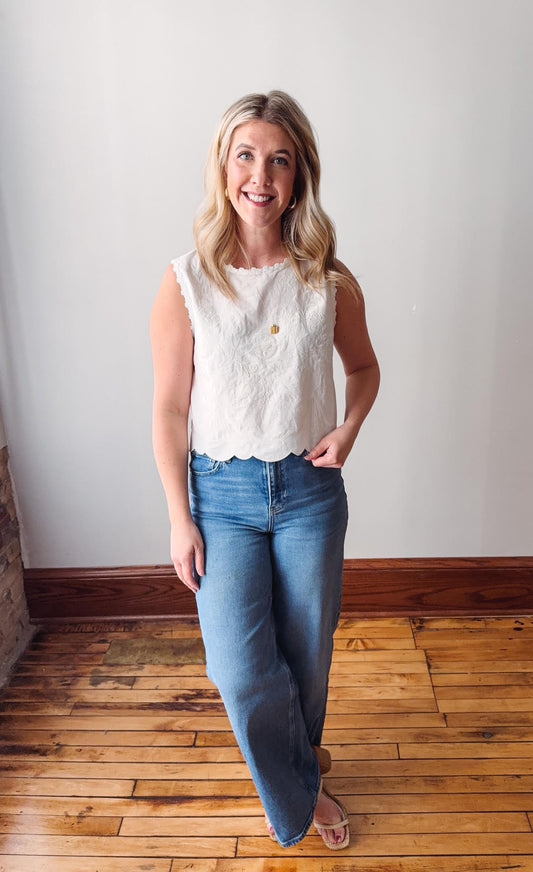 Woman wearing a white sleeveless top and blue jeans standing on a wooden floor.