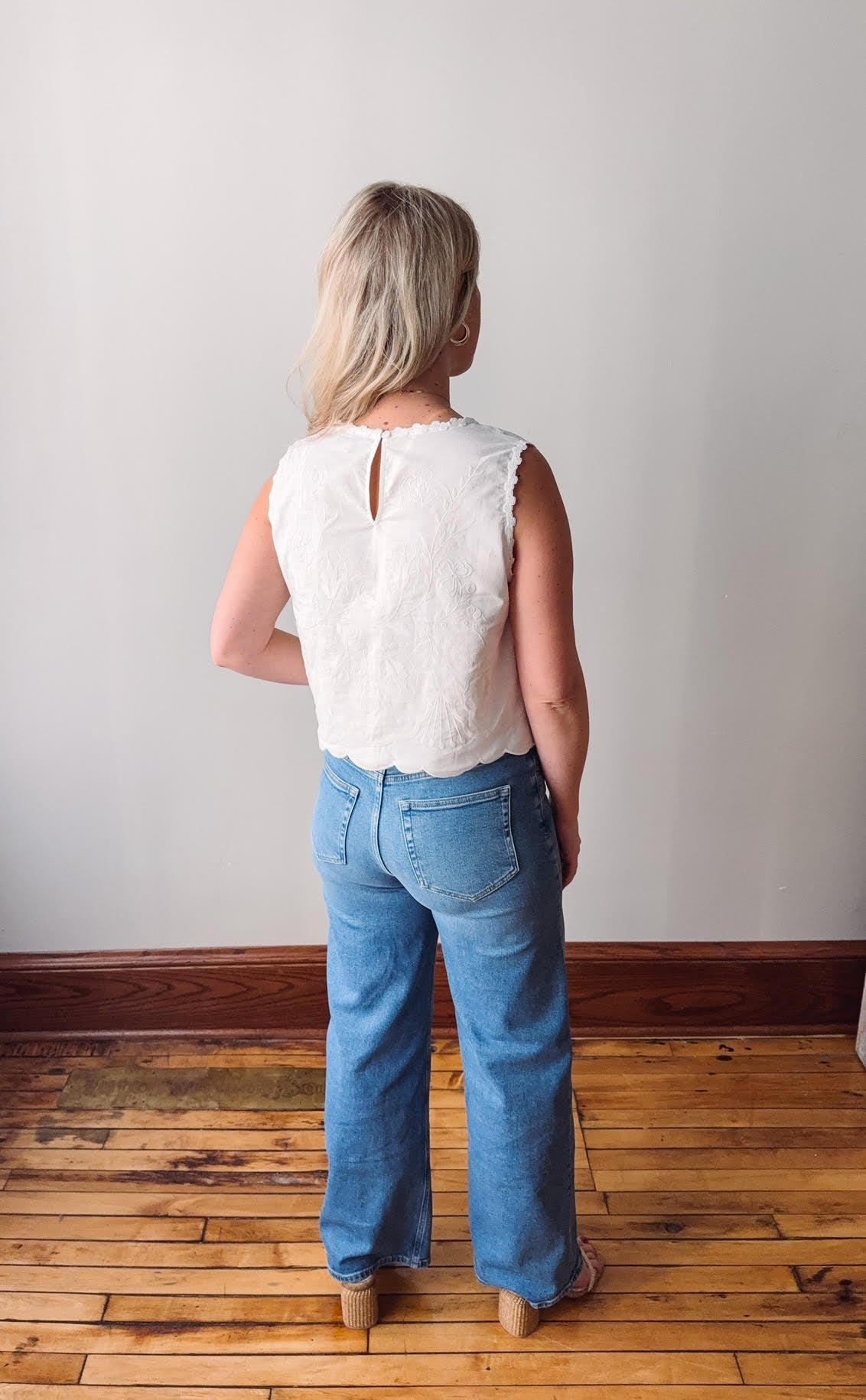 Person wearing a white sleeveless top and blue jeans standing in a room with wooden flooring and a plain wall.