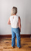 Person wearing a white sleeveless top and blue jeans standing in a room with wooden flooring and a plain wall.