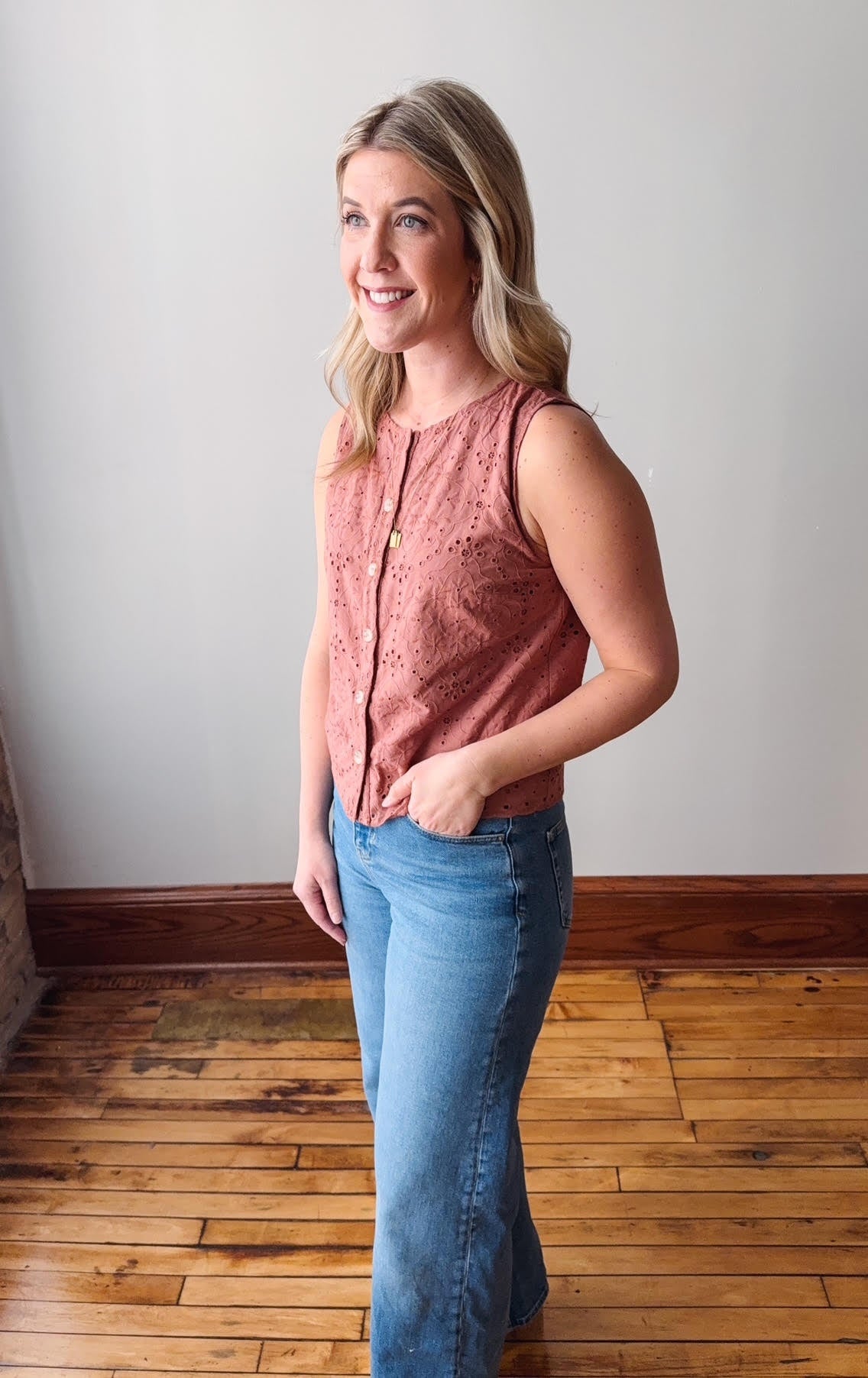 Woman wearing a pink sleeveless top and blue jeans standing in a room with wooden flooring.